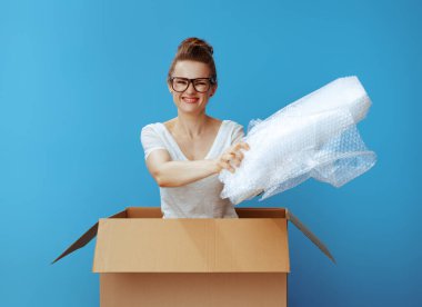 smiling young woman in white t-shirt in a cardboard box giving air bubble film packaging material isolated on blue clipart