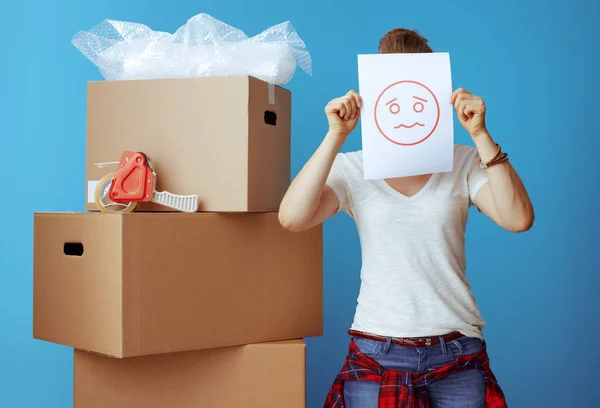 modern woman in white t-shirt near cardboard box hiding behind a sheet of paper with unhappy smiley isolated on blue