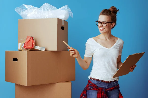 微笑的现代妇女在白色 T恤附近纸板箱使用移动的清单和计数箱子在蓝色背景 — 图库照片