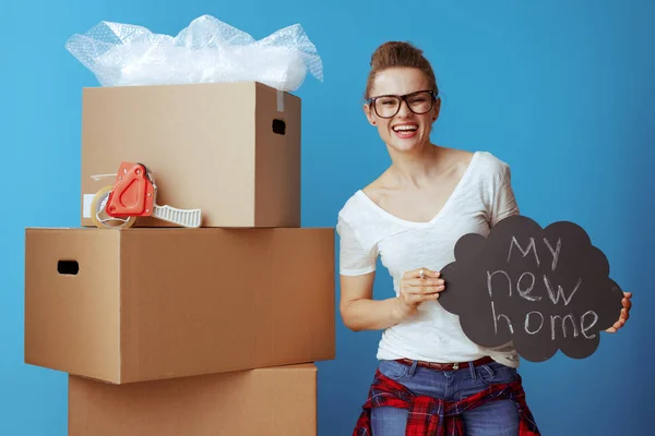 青の背景に私の新しい家のボードを表示段ボール箱の近く白い シャツで幸せな現代の女性 — ストック写真