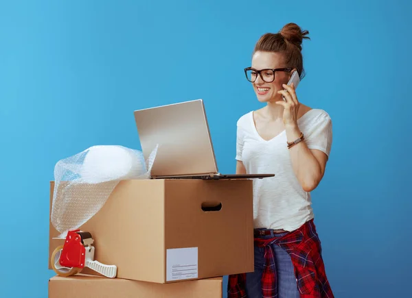 微笑的年轻女子在白色 T恤附近的纸板箱看着笔记本电脑屏幕 并在蓝色背景下的手机交谈 — 图库照片