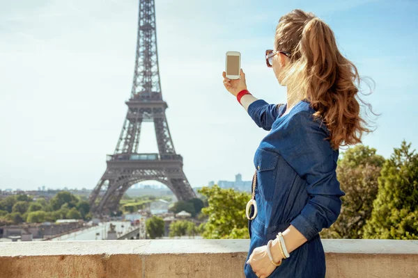 从后面看到穿着蓝色牛仔裤的时尚女子与智能手机在法国巴黎的埃菲尔铁塔合影 — 图库照片