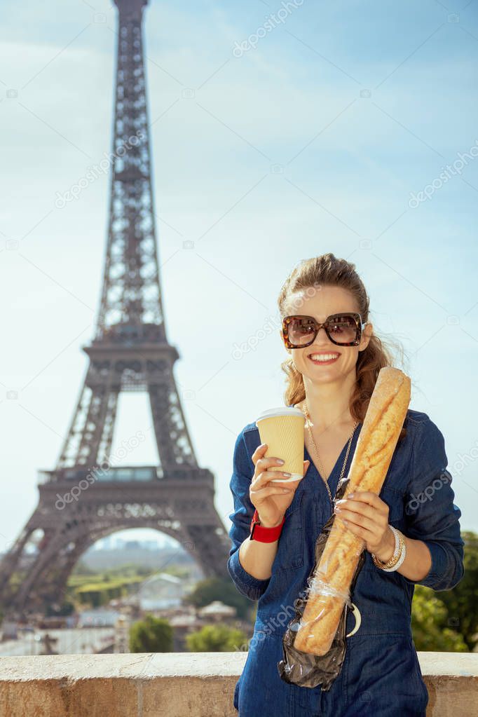 Portrait of smiling stylish tourist woman in blue jeans overall with coffee cup and baguette having excursion not far from Eiffel tower in Paris, France.