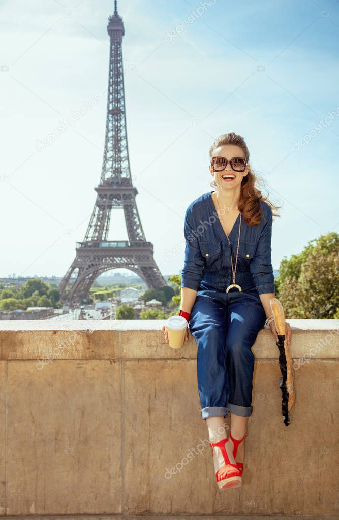 Portrait of happy trendy tourist woman in blue jeans overall with coffee cup and baguette having excursion against clear view of the Eiffel Tower in Paris, France.