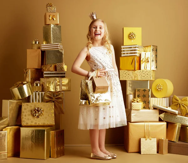 在一面普通的墙前 一个金色的礼物中的2堆金色礼物中 面带微笑的现代小公主的全长肖像 穿着米色的礼服和闪光灯 头上戴着金色的购物袋 — 图库照片