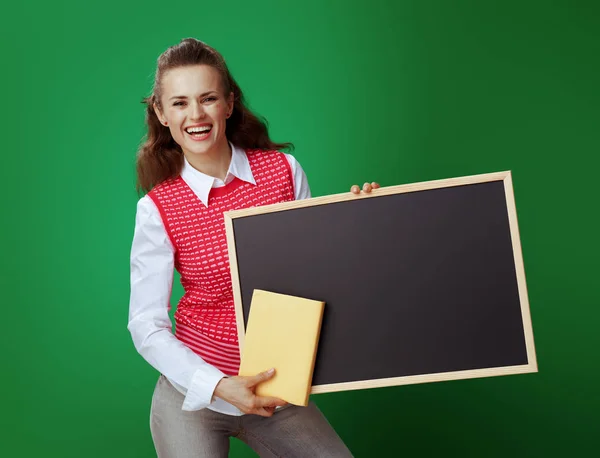 Sorrindo mulher estudante ajuste com livro amarelo mostrando quadro negro — Fotografia de Stock