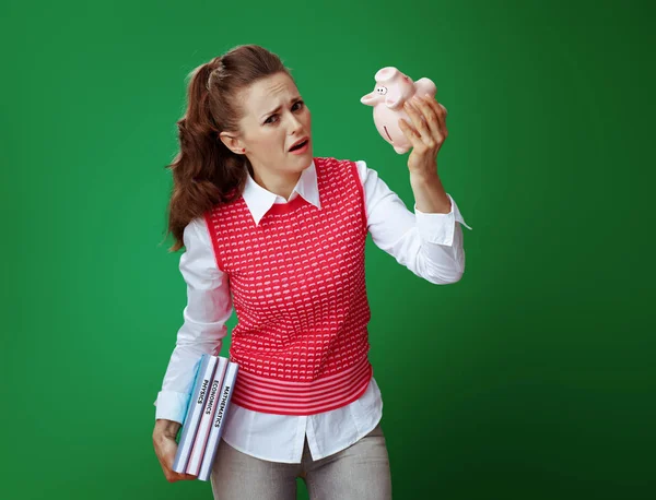 Unhappy student woman with textbooks shaking pink piggy bank — Stock Photo, Image