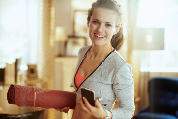 Mujer con colchoneta de fitness y teléfono inteligente en la sala de estar moderna —  Fotos de Stock