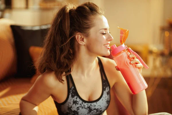 モダンなリビング ルームに補足蛋白質とシェーカーから飲むスポーツ服で健康な女性の笑みを浮かべて — ストック写真