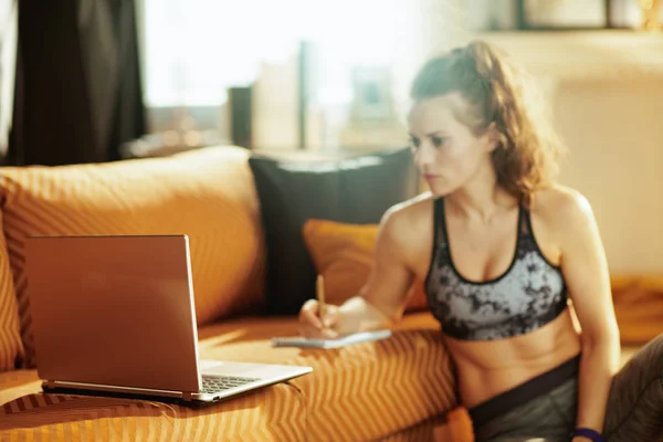 特写镜头在笔记本电脑和活跃的妇女在运动服与记事本和笔做笔记 同时在现代家庭的背景下在互联网上观看健身教程 — 图库照片