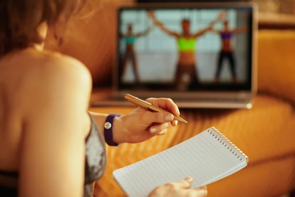 メモ帳とペンを現代の家でオンライン ストリーミング サイト内のノート パソコンを使用しながらメモを取るスポーツ服の若いスポーツ女性のクローズ アップ — ストック写真