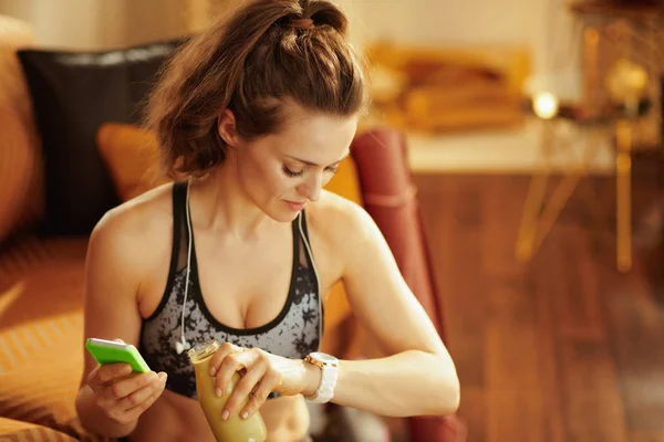 适合运动女性在健身服装与耳机和一瓶绿色冰沙使用智能手表跟踪心率在现代房子的健身应用程序 — 图库照片