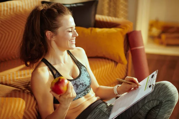 happy active woman in sport clothes with an apple and clipboard filling meal plan at modern home.
