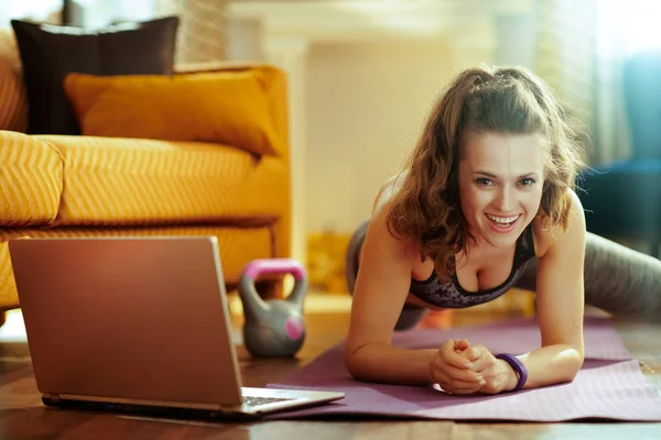 微笑适合妇女在现代房子的运动服装使用在线流媒体健身网站在笔记本电脑和做有氧运动在健身垫 — 图库照片