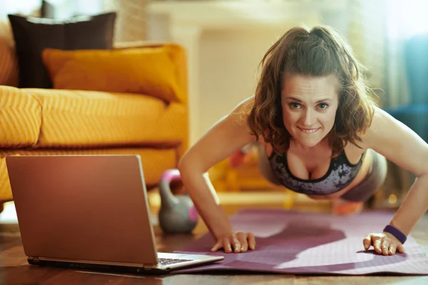 active woman in fitness clothes in the modern house using laptop to watch fitness streaming on internet and doing pushups on fitness mat.