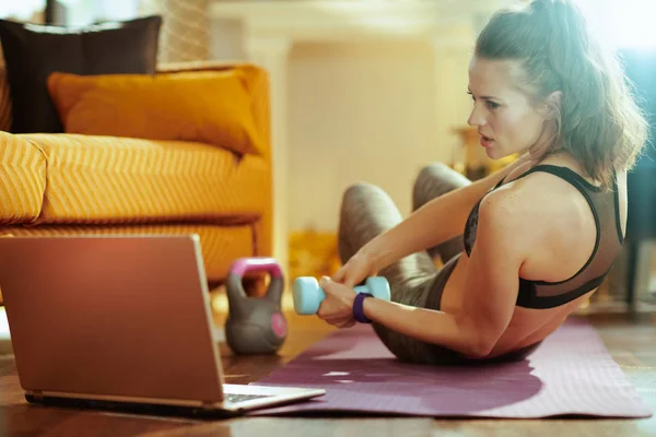healthy woman in fitness clothes using online streaming fitness site in laptop and doing exercises with dumbbells on fitness mat in the modern house.