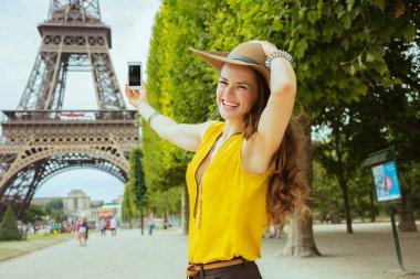 sarı bluz ve şapka gülümseyen trendy yalnız gezgin kadın Paris'te Eyfel kulesiönünde cep telefonu ile fotoğraf çekmek, Fransa.