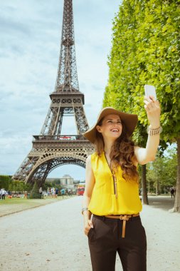 sarı bluz ve şapka mutlu modern yalnız gezgin kadın Paris'te Eyfel kulesine karşı akıllı telefon ile selfie alarak, Fransa.