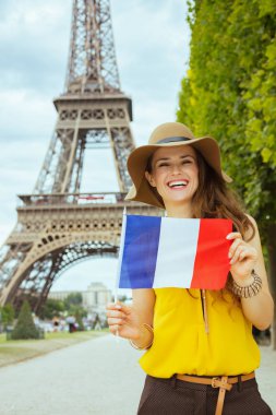 Paris'te Eyfel Kulesi'nin net görünümüne karşı Fransız bayrağı gösteren sarı bluz ve şapka gülümseyen trendy turist kadın, Fransa.