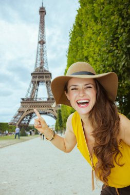 Champ de Mars'ta sarı bluz ve şapka ile gülümseyen genç turist kadın Paris'te Eyfel kulesini işaret eden, Fransa