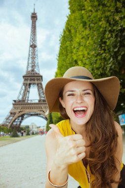 Paris'te Eyfel kulesinin önünde sarı bluz ve şapka mutlu genç yalnız turist kadın, Fransa kadar başparmak gösteren.