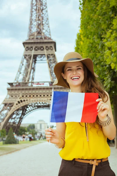 smiling trendy tourist woman in yellow blouse and hat showing French flag against clear view of the Eiffel Tower in Paris, France.