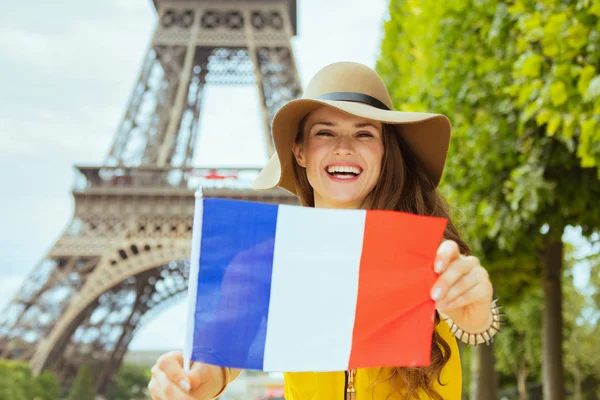 happy young solo traveller woman in yellow blouse and hat showing French flag against clear view of the Eiffel Tower in Paris, France.