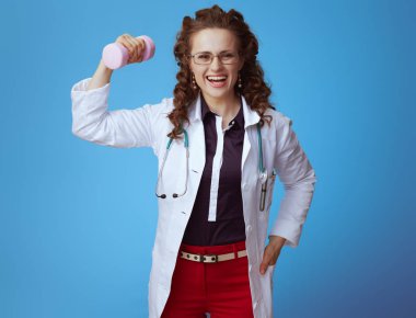 bue gömlek, kırmızı pantolon ve pembe dambıl ile beyaz tıbbi bornoz gülümseyen zarif doktor kadın mavi arka plan üzerinde izole pazı gösteren.