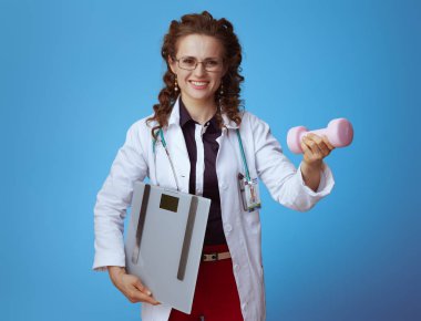 bue gömlek mutlu zarif tıp uygulayıcısı kadın, kırmızı pantolon ve ağırlık ölçeği ile beyaz tıbbi bornoz mavi arka plan üzerinde dambıl veren.