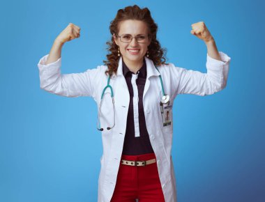 bue gömlek, kırmızı pantolon ve beyaz tıbbi bornoz mavi izole pazı gösteren gülümseyen modern hekim kadın.