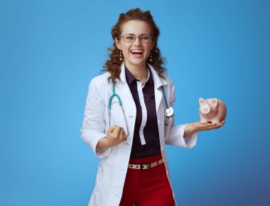 bue gömlek mutlu modern tıp uygulayıcısı kadın, kırmızı pantolon ve kumbara ile beyaz tıbbi cüppe mavi arka plan karşı seviniyor.