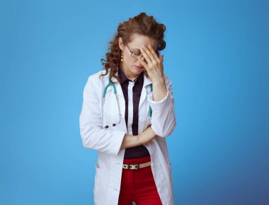 Bue gömlek yorgun modern doktor kadın Portresi, kırmızı pantolon ve mavi arka plan izole beyaz tıbbi bornoz.