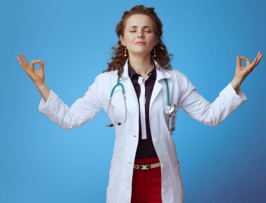 Bue gömlek rahat modern hekim kadın Portresi, kırmızı pantolon ve beyaz tıbbi bornoz mavi arka plan üzerinde yoga yapıyor.