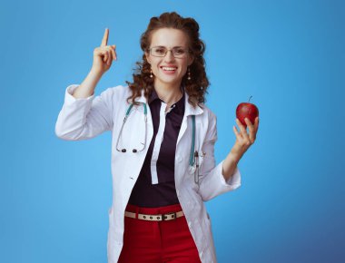 bue gömlek, kırmızı pantolon ve beyaz tıbbi bornoz bir elma gösteren gülümseyen modern hekim kadın mavi izole fikir var.