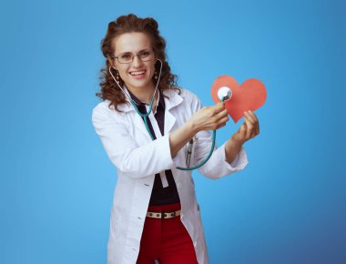 bue gömlek, kırmızı pantolon ve beyaz tıbbi bornoz stetoskop kırmızı kağıt kalp mavi izole ile dinleme gülümseyen modern hekim kadın.