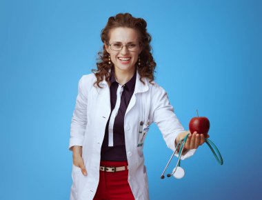 bue gömlek mutlu modern tıp uygulayıcısı kadın, kırmızı pantolon ve beyaz tıbbi bornoz mavi arka plan üzerinde stetoskop ve kırmızı elma veren.