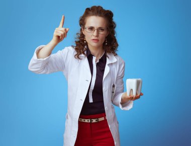 bue gömlek modern doktor kadın, kırmızı pantolon ve beyaz tıbbi bornoz beyaz diş uyarı ile yükselir parmak mavi izole.