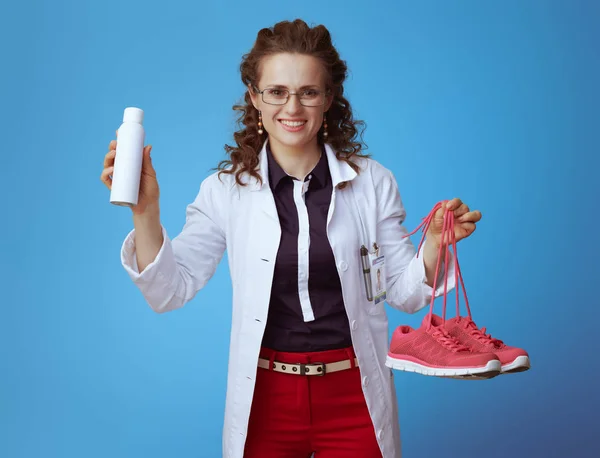 快乐优雅的医生妇女在Bue衬衫 红色裤子和白色医疗长袍显示健身运动鞋和鞋除臭剂喷雾隔离在蓝色背景 — 图库照片