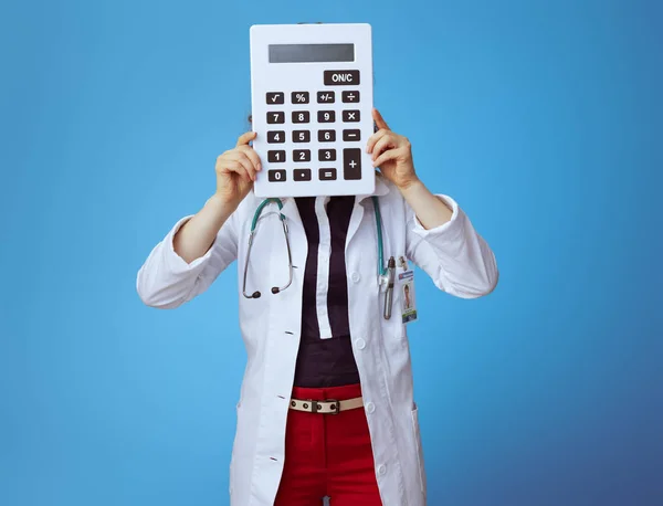 优雅的医生女人在Bue衬衫 红色裤子和白色医疗长袍拿着大白色计算器在前面的脸隔离在蓝色 — 图库照片