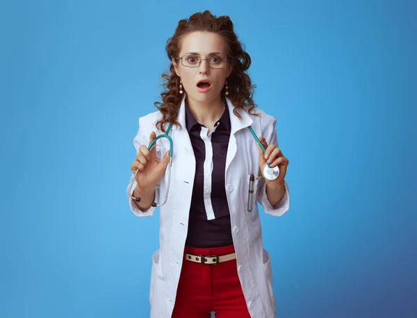 ブエシャツ 赤いパンツと青い背景に隔離された白い医療ローブで驚いたエレガントな医師の女性の肖像画 — ストック写真