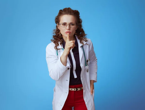 Portrait of elegant medical practitioner woman in bue shirt, red pants and white medical robe showing shh gesture against blue background.