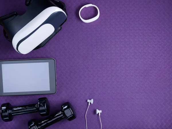 Closeup on VR glasses, white fitness tracker, headphones, tablet PC and black dumbbells laying on purple fitness mat. Flat Lay.