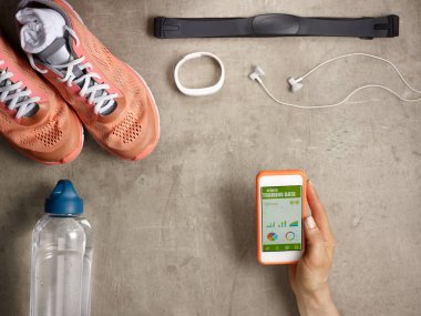 Spor ayakkabıları, kalp atış hızı monitörü, bir şişe su, beyaz fitness izci, yerde yatan kulaklıklar ve spor eğitim verileri ile akıllı telefon tutan kadın el üzerinde closeup
