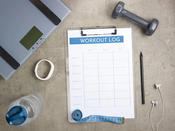 Closeup on laying on the floor weight scales, grey dumbbell, white fitness tracker, bottle of water, tape measure, headphones, black pen and workout log on clipboard.