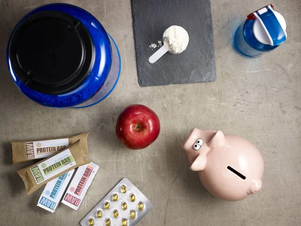 Closeup on big blue protein jar, shaker, measuring spoon with powder, raw protein bars, red apple, sport supplements in tablets and piggy bank laying on the floor.