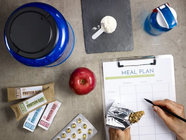 Closeup on protein jar, shaker, measuring spoon, raw protein bars, red apple, sport supplements in tablets laying on floor and female hand with pen filling meal plan and holding fitness energy bar.