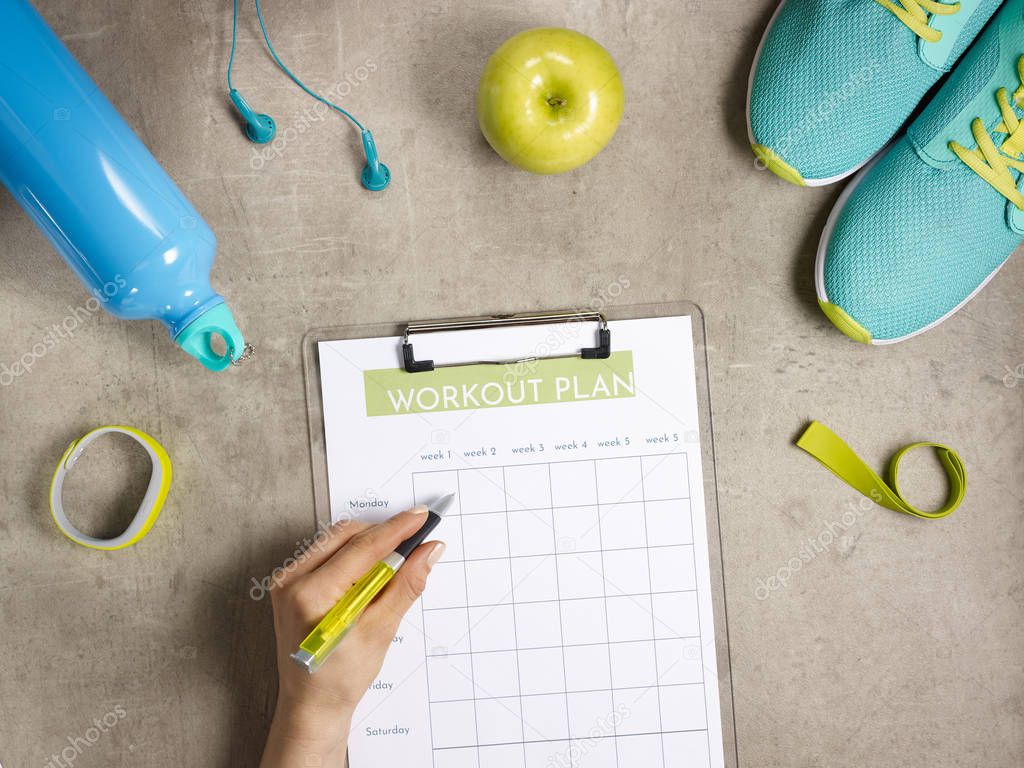 Closeup on green apple, headphones, elastic band, sneakers, fitness tracker, bottle of water laying on the floor and female hand filling workout plan on clipboard with pen.