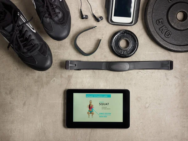 Closeup on black sneakers, headphones, fitness tracker, hear rate monitor, smartphone in running armband, bottle of water, weight plate and tablet PC with home fitness app laying on the floor.