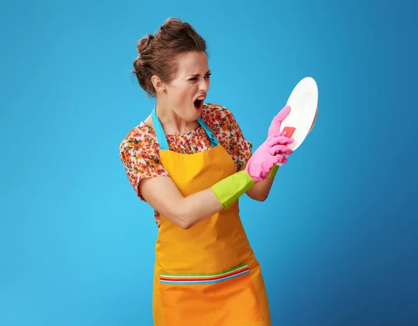 Angry young woman with sponge washing dish on blue — Stock Photo, Image