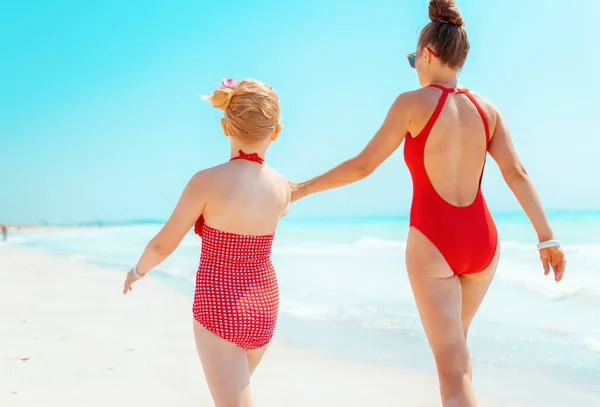 Young mother and daughter in red swimwear on seashore walking — Stock Photo, Image